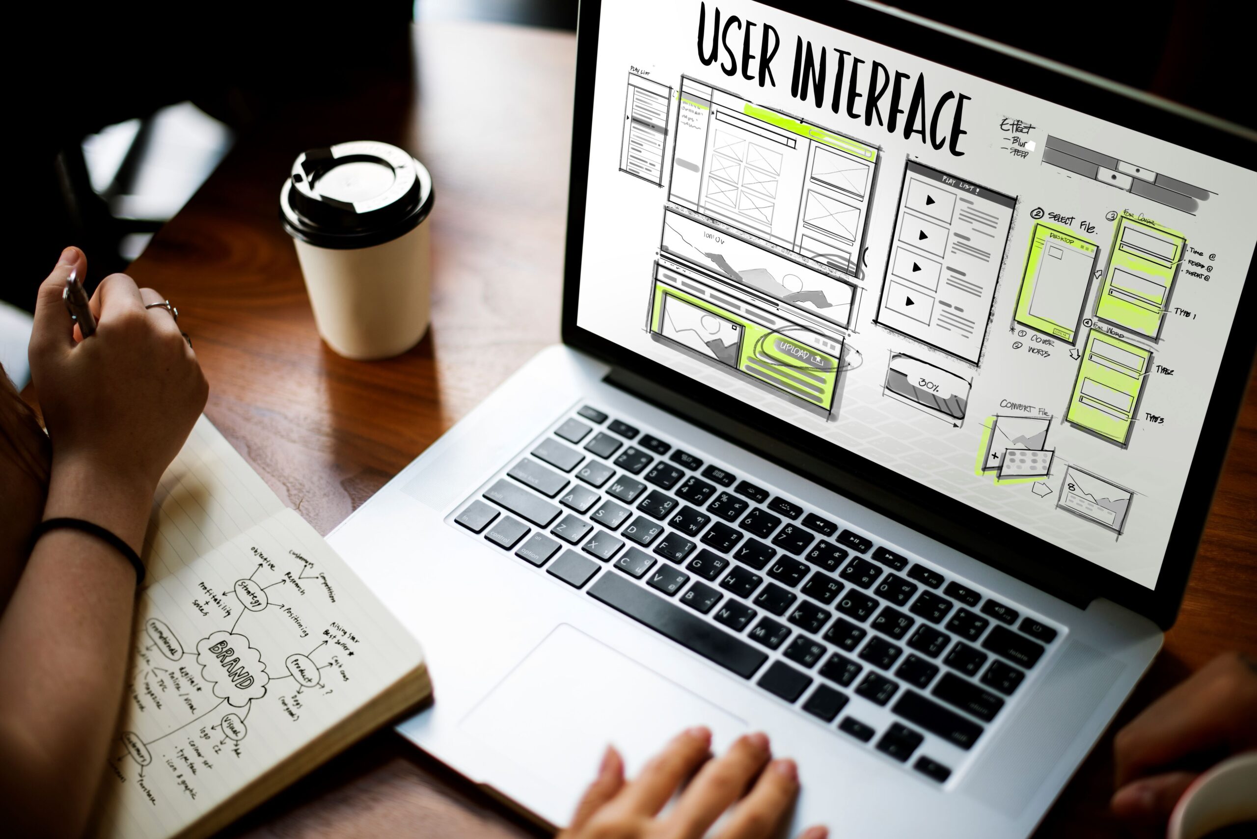 Laptop displaying user interface design sketches, with a hand taking notes beside a coffee cup, representing website development and design services.