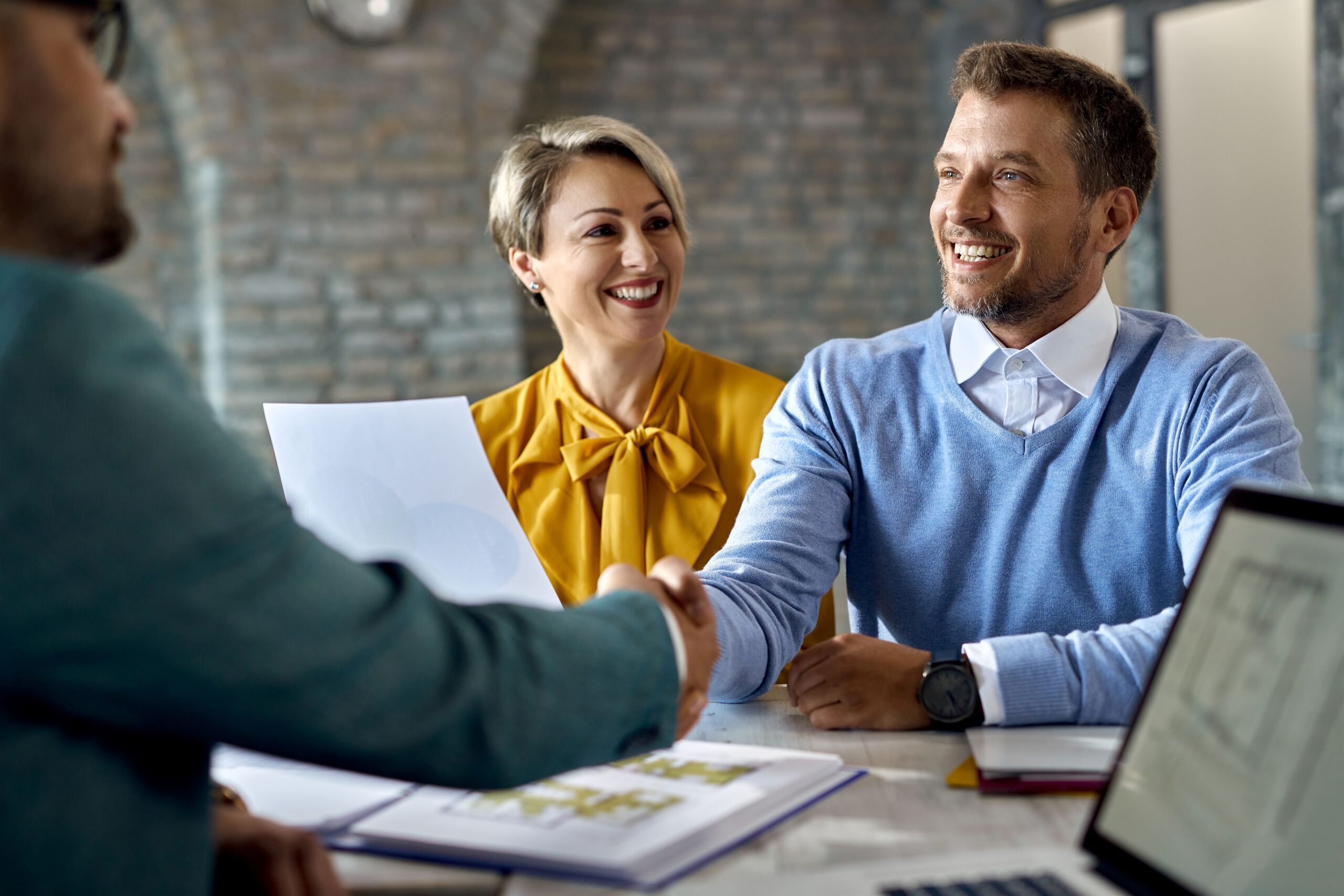Business meeting with a man and woman shaking hands, smiling, discussing marketing strategies, with documents and laptops on the table, emphasizing collaboration and client engagement.