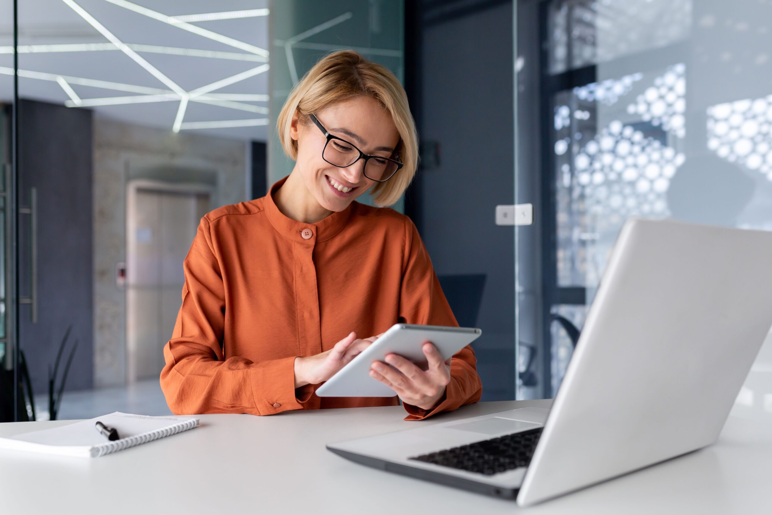 Young businesswoman in an orange blouse using a tablet at a modern office desk, with a laptop and notepad, illustrating the importance of digital presence for entrepreneurs.