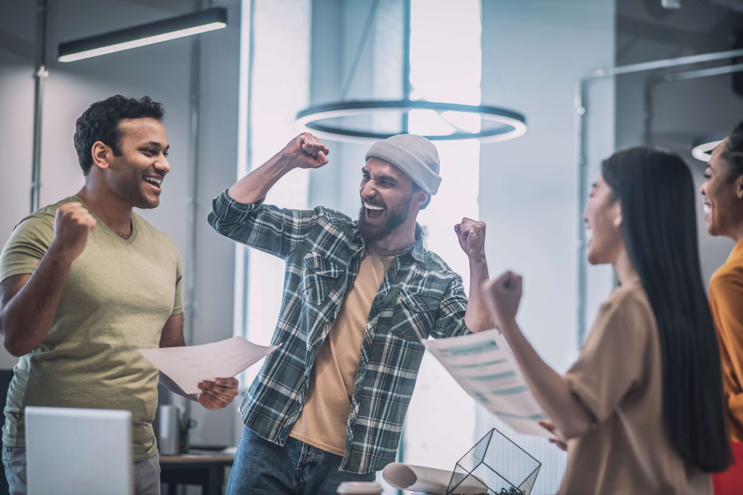 Group of diverse young professionals celebrating success in a modern office, with raised fists and smiles, holding documents related to digital marketing and business growth.